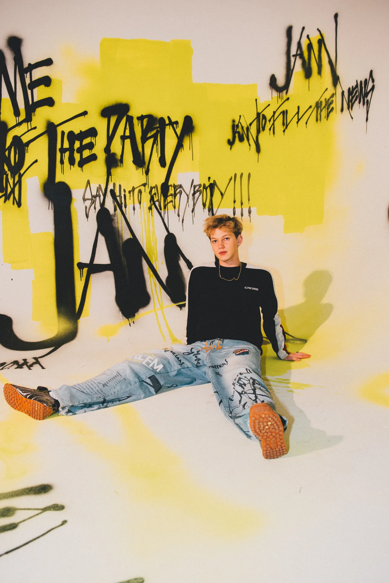 Artysta Jann siedzący na podłodze, swobodnie pozujący przed ścianą pokrytą żółto-czarnym graffiti 'WELCOME TO THE PARTY JANN' podczas eventu Jann Popup Store.