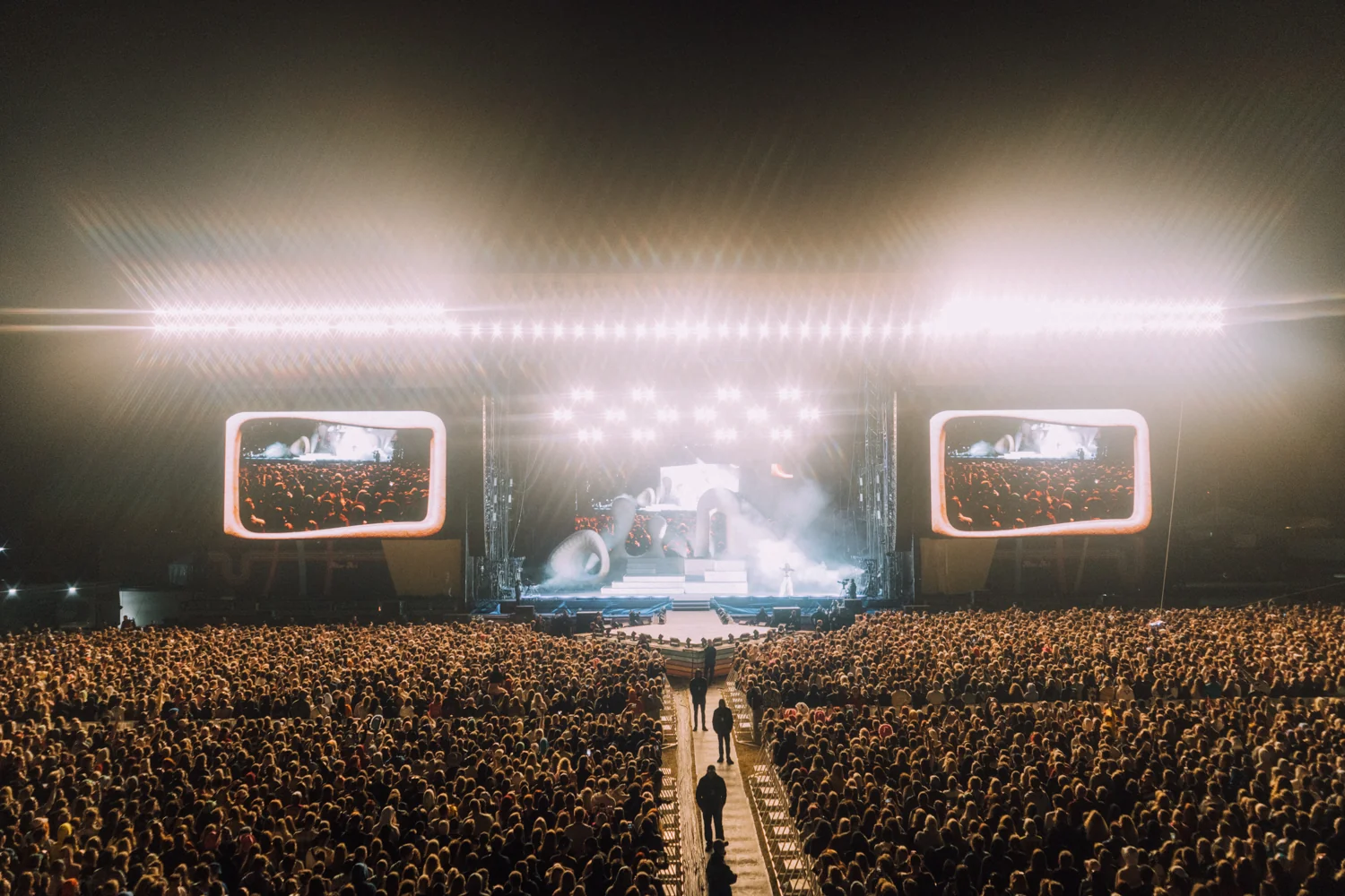 Szerokie ujęcie ogromnej publiczności podczas nocnego koncertu festiwalowego, z jasno oświetloną sceną i ekranami LED.