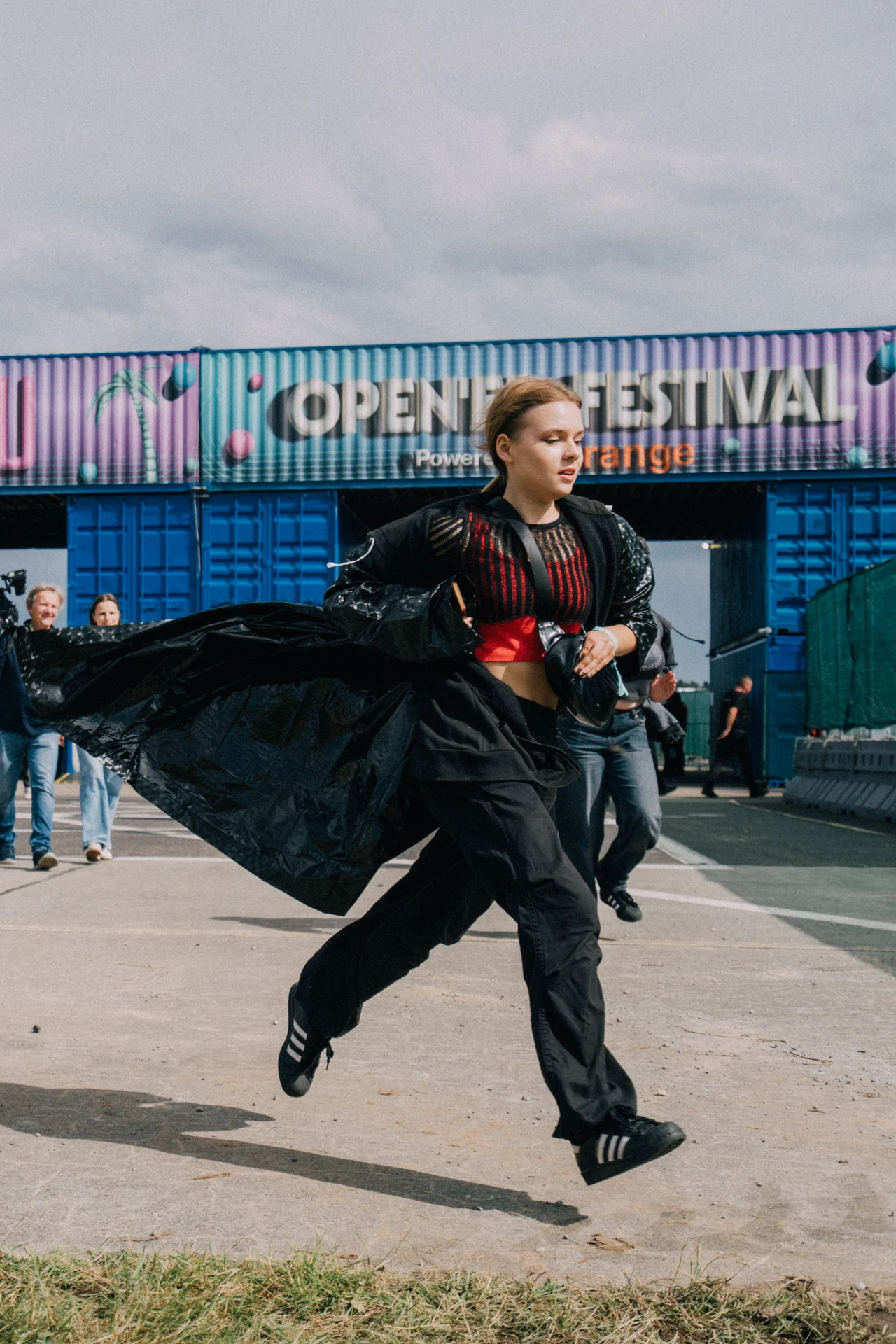 Kobieta w czarnej stylizacji biegnąca przed kolorową ścianą kontenerów z brandingiem 'OPEN'ER FESTIVAL', na terenie festiwalu.
