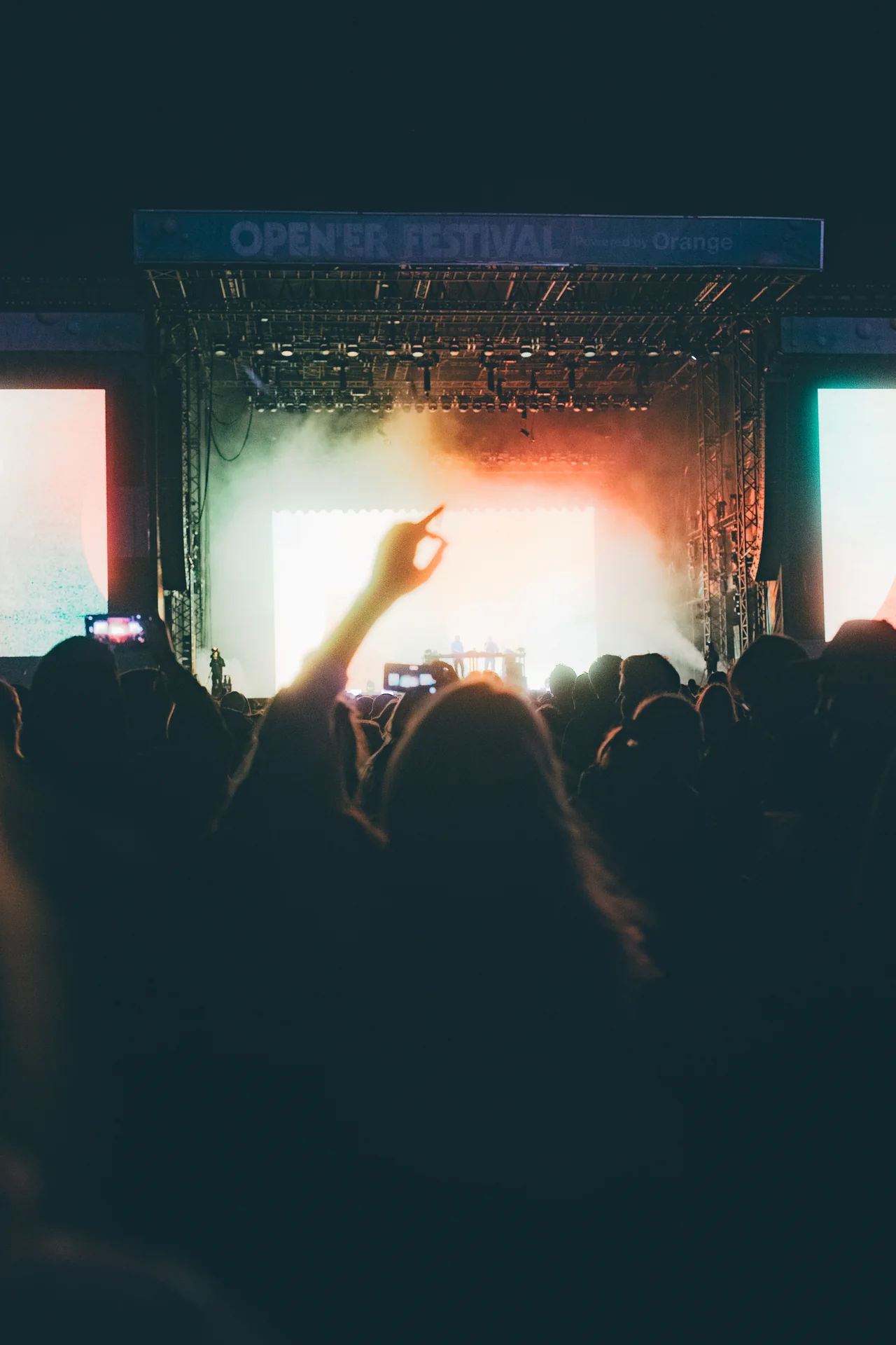 Sylwetka uniesionej dłoni w kierunku jasno oświetlonej sceny Opener Festival 2024, z zespołem i publicznością w tle