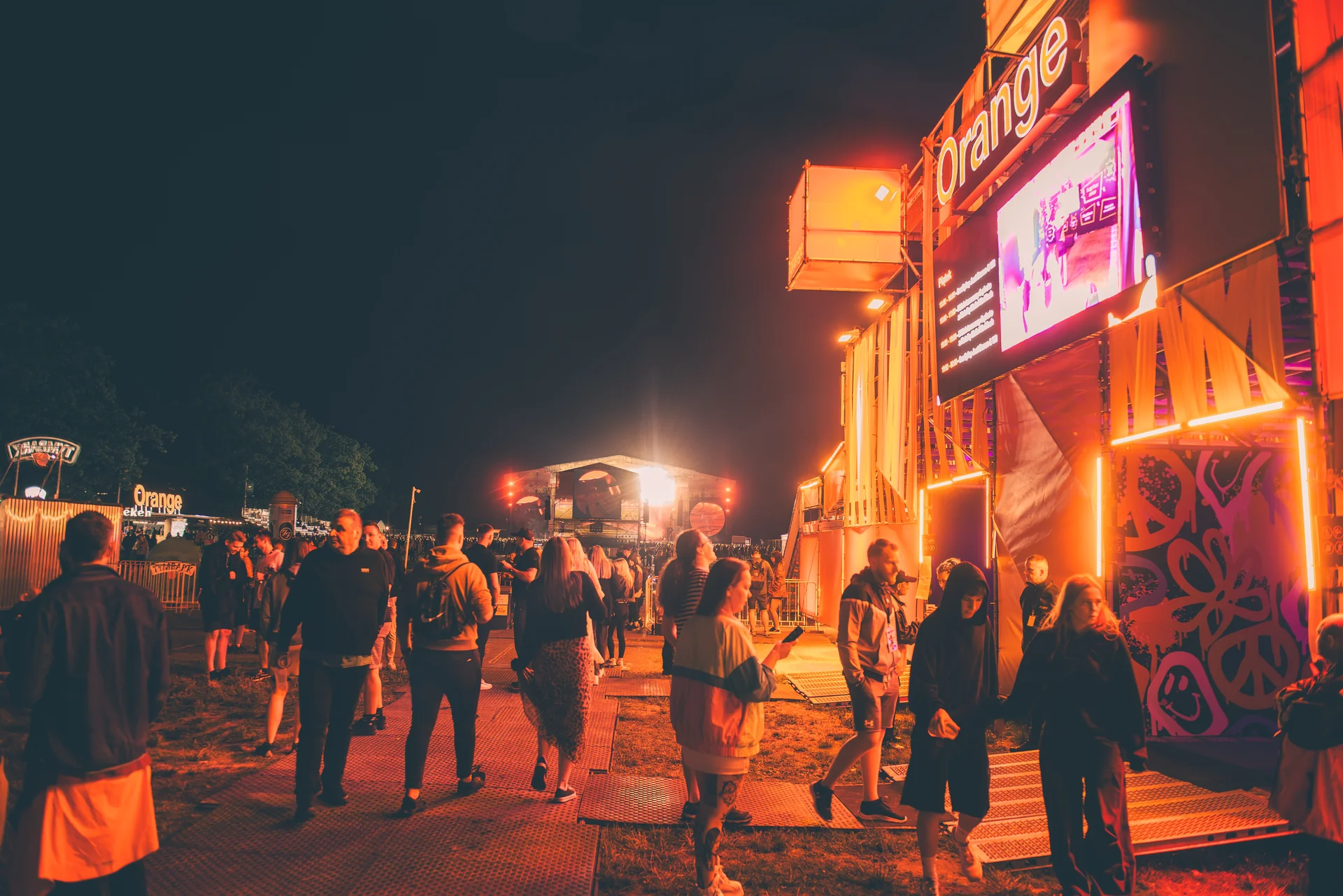 Ludzie spacerujący po terenie Orange Warsaw Festival 2024 w nocy, z podświetlonymi stoiskami i dużym logo 'Orange' w tle.