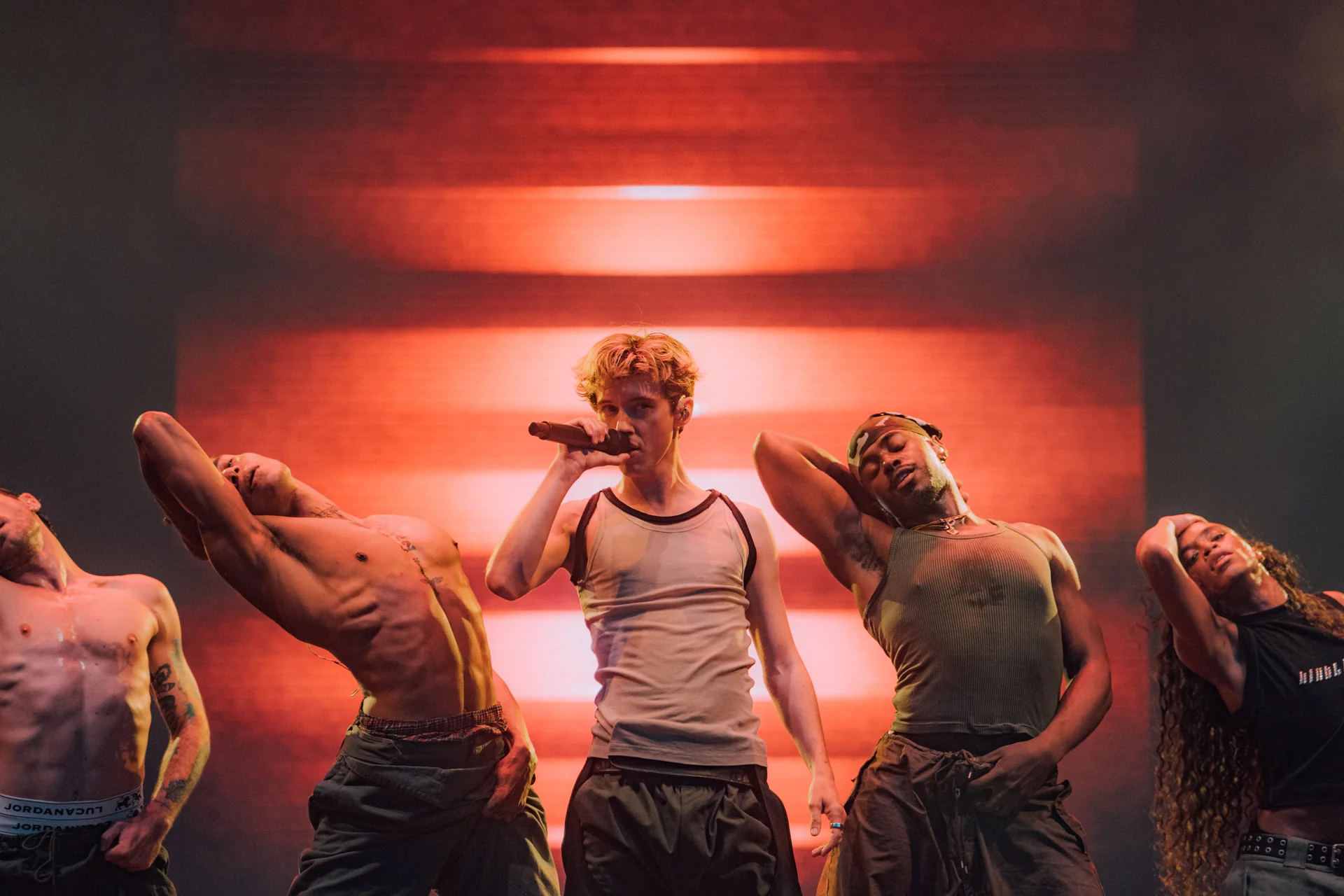 Troye Sivan w białym topie i spodniach, śpiewający do mikrofonu otoczony półnagimi tancerzami, na scenie Orange Warsaw Festival 2024 w czerwonym świetle.
