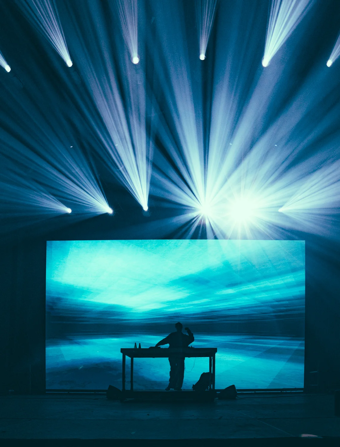 Sylwetka DJ-a na scenie Orange Warsaw Festiwal 2025, stojącego przed dużym ekranem LED z niebieskimi wizualizacjami i promieniami światła.