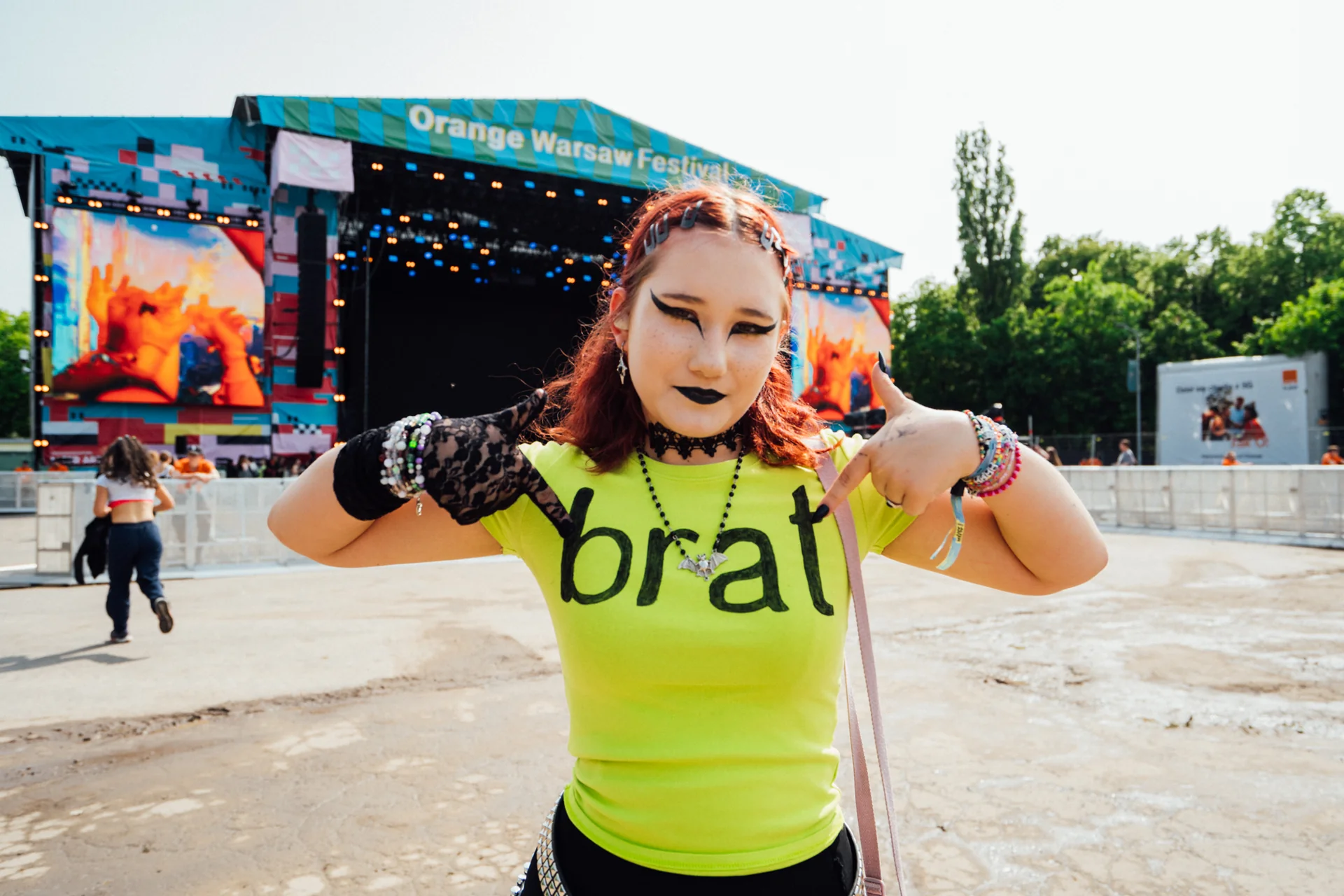 Młoda kobieta z rudymi włosami i wyrazistym makijażem, w neonowej koszulce z napisem 'brat', pozująca na Orange Warsaw Festiwal 2025.