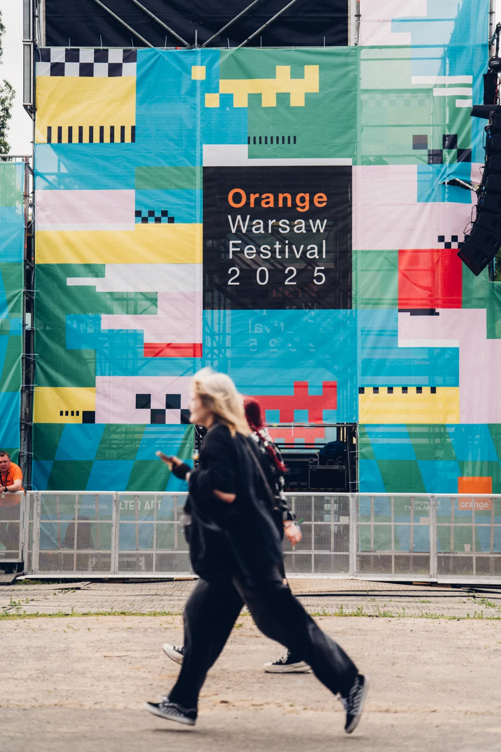 Ludzie spacerujący po terenie Orange Warsaw Festiwal 2025, z dużym kolorowym banerem reklamującym edycję 2025 festiwalu w tle.
