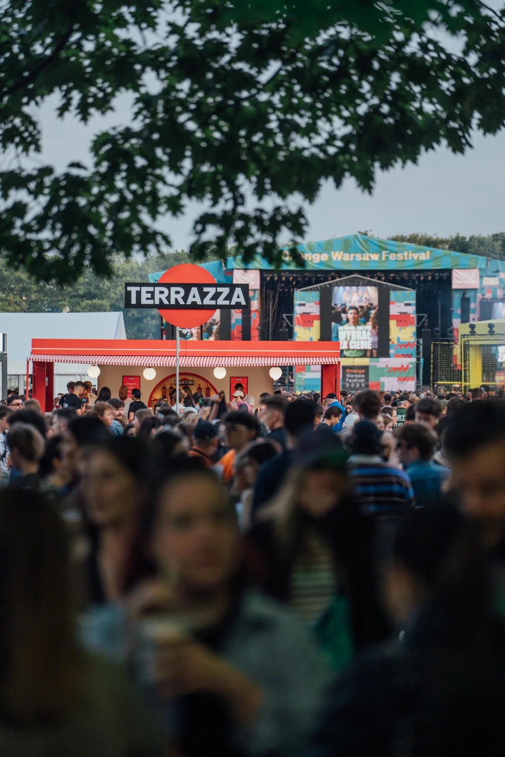 Tłum ludzi na Orange Warsaw Festiwal 2025 przed stoiskiem 'Terrazza', z fragmentem głównej sceny festiwalu w tle, w świetle dziennym.