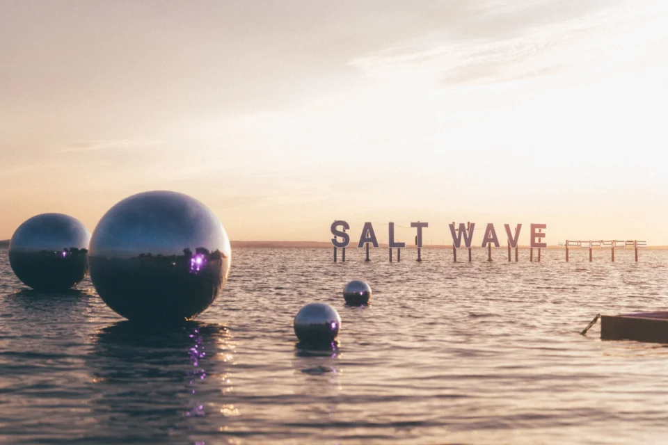 Metalowe kule unoszące się na wodzie, z napisem 'SALT WAVE' na horyzoncie, tworzące artystyczną instalację na festiwalu Salt Wave 2024.