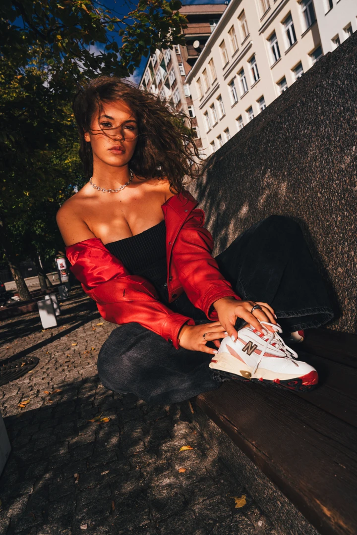 Alicja Szemplinska pochylona w czerwonej kurtce i czarnych spodniach eksponuje buty New Balance 1000 w miejskim wnętrzu