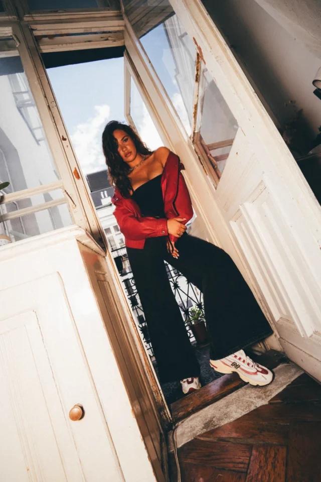 Alicja Szemplinska stoi w otwartych drzwiach balkonu w czerwonej kurtce, czarnych spodniach i białych butach New Balance 1000