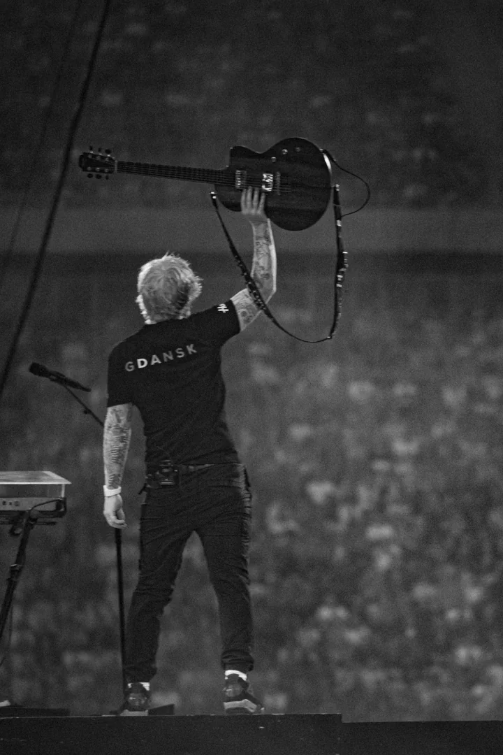 Muzyk z tatuażami unosi gitarę w geście triumfu, stojąc na scenie przed tłumem fanów