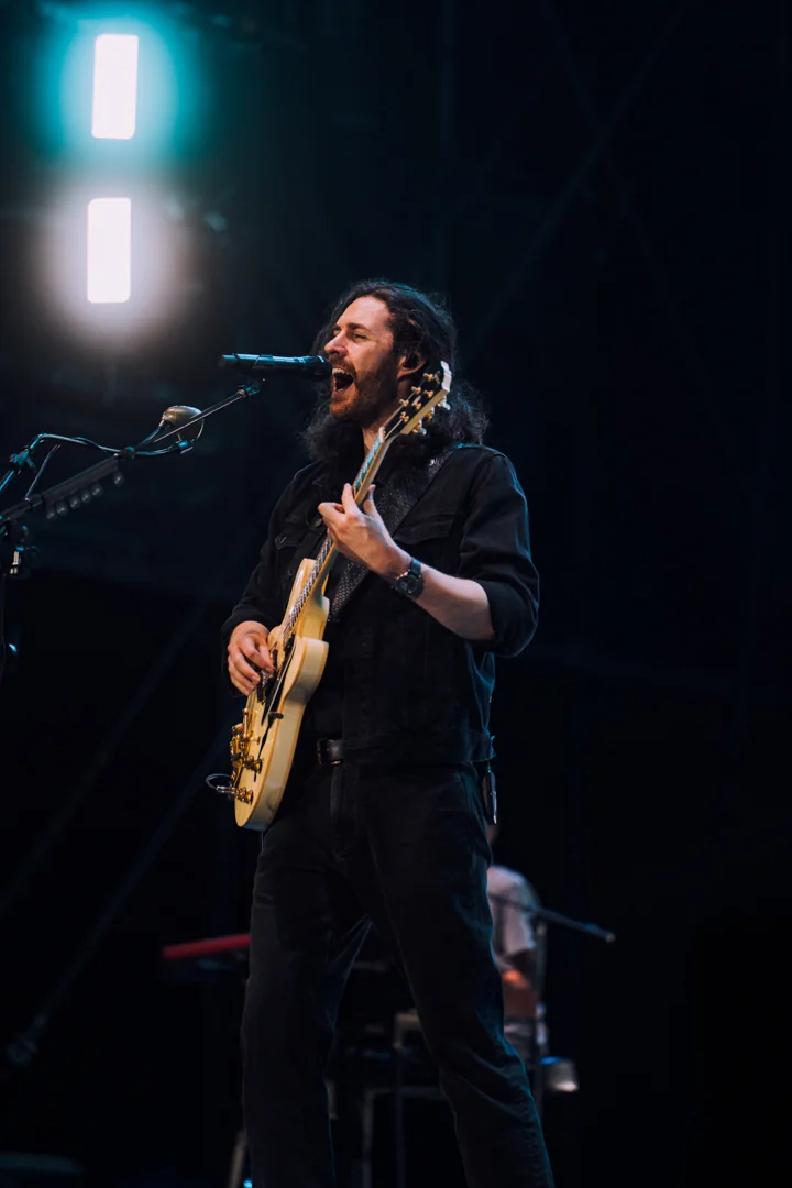 Muzyk z długimi włosami gra na jasnej gitarze, śpiewając z pasją przy intensywnym świetle sceny
