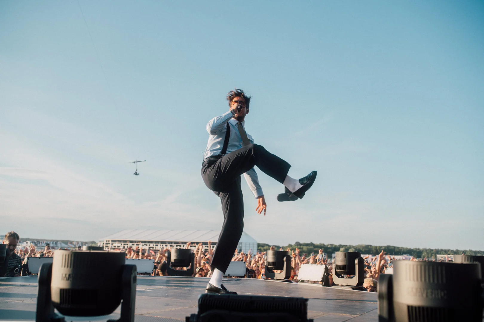 Mężczyzna w białej koszuli i czarnych spodniach energicznie śpiewa, podnosząc nogę na scenie przed tłumem
